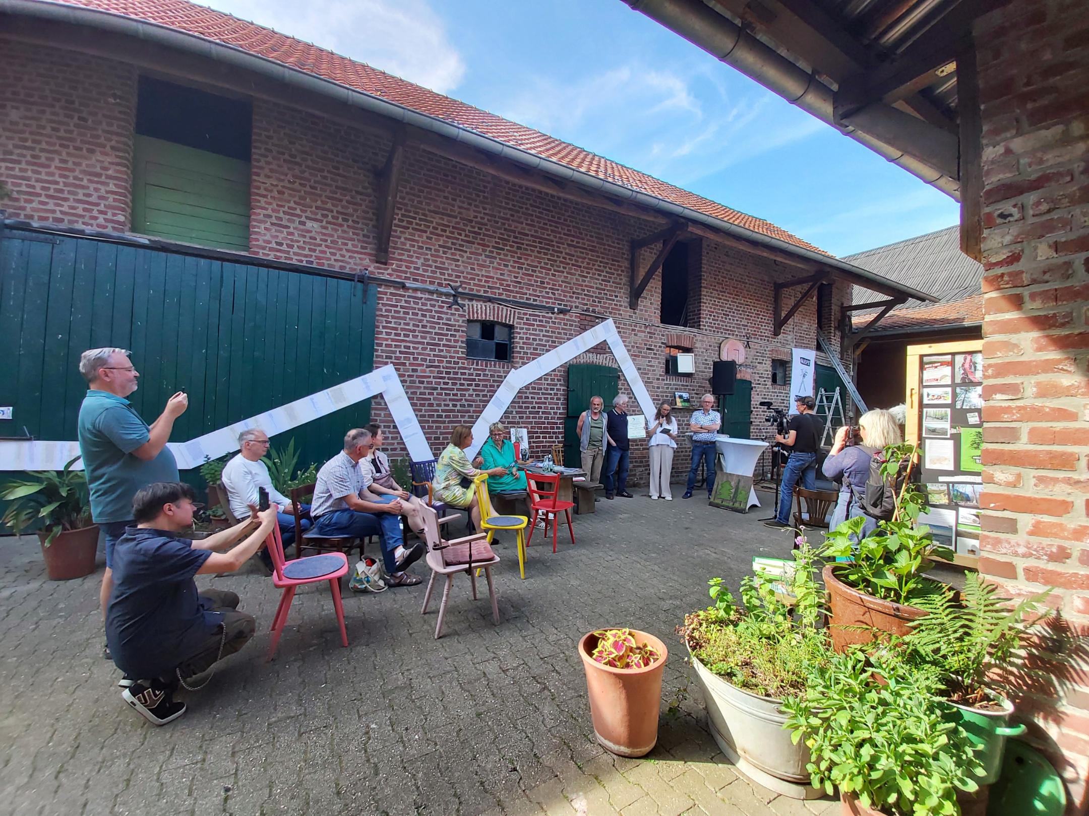 Menschen sitzen und stehen in einem Innenhof mit Backsteinmauern, einige machen Fotos, andere hören einer Person zu, die vor einem Mikrofon spricht.