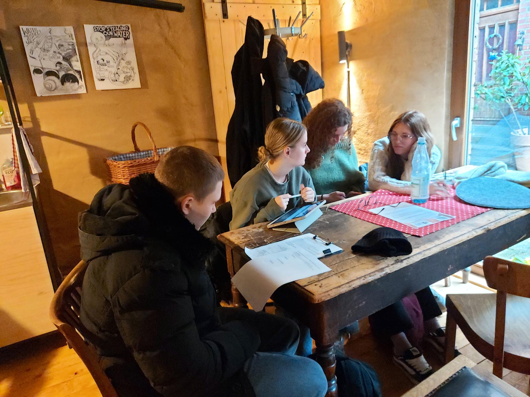 Vier Personen sitzen an einem rustikalen Holztisch mit Papieren und einem Tablet in einem gemütlichen Raum mit Holzvertäfelung und Garderobe im Hintergrund