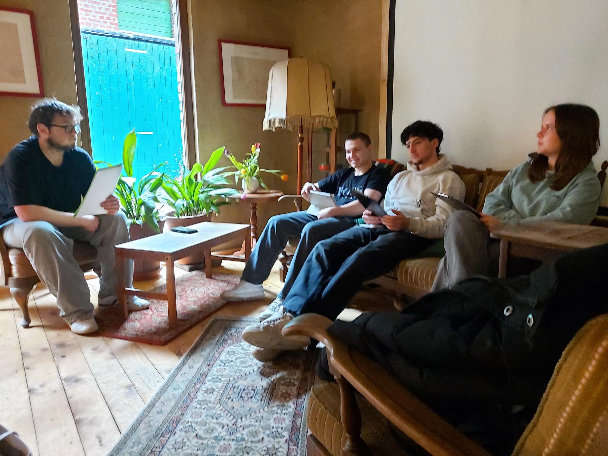 Vier Personen sitzen in einem Wohnzimmer auf Sesseln und einem Sofa, drei halten Tablets oder Smartphones, ein Tisch mit Pflanzen steht in der Mitte.