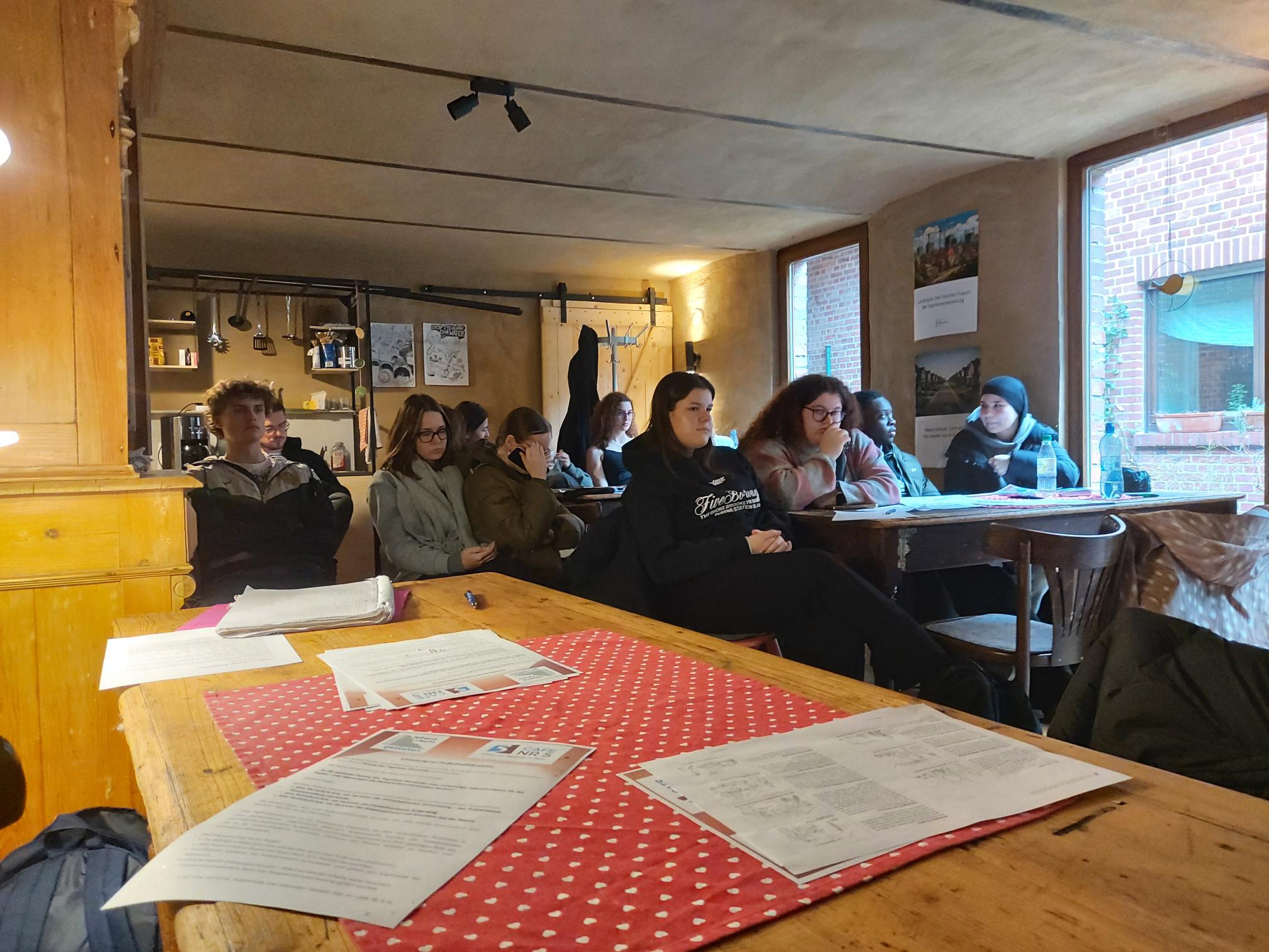 Mehrere Personen sitzen an Holztischen in einem Raum mit großen Fenstern und Deckenlampen, auf den Tischen liegen Papiere und ein rot-weiß gepunktetes Tischtuch.