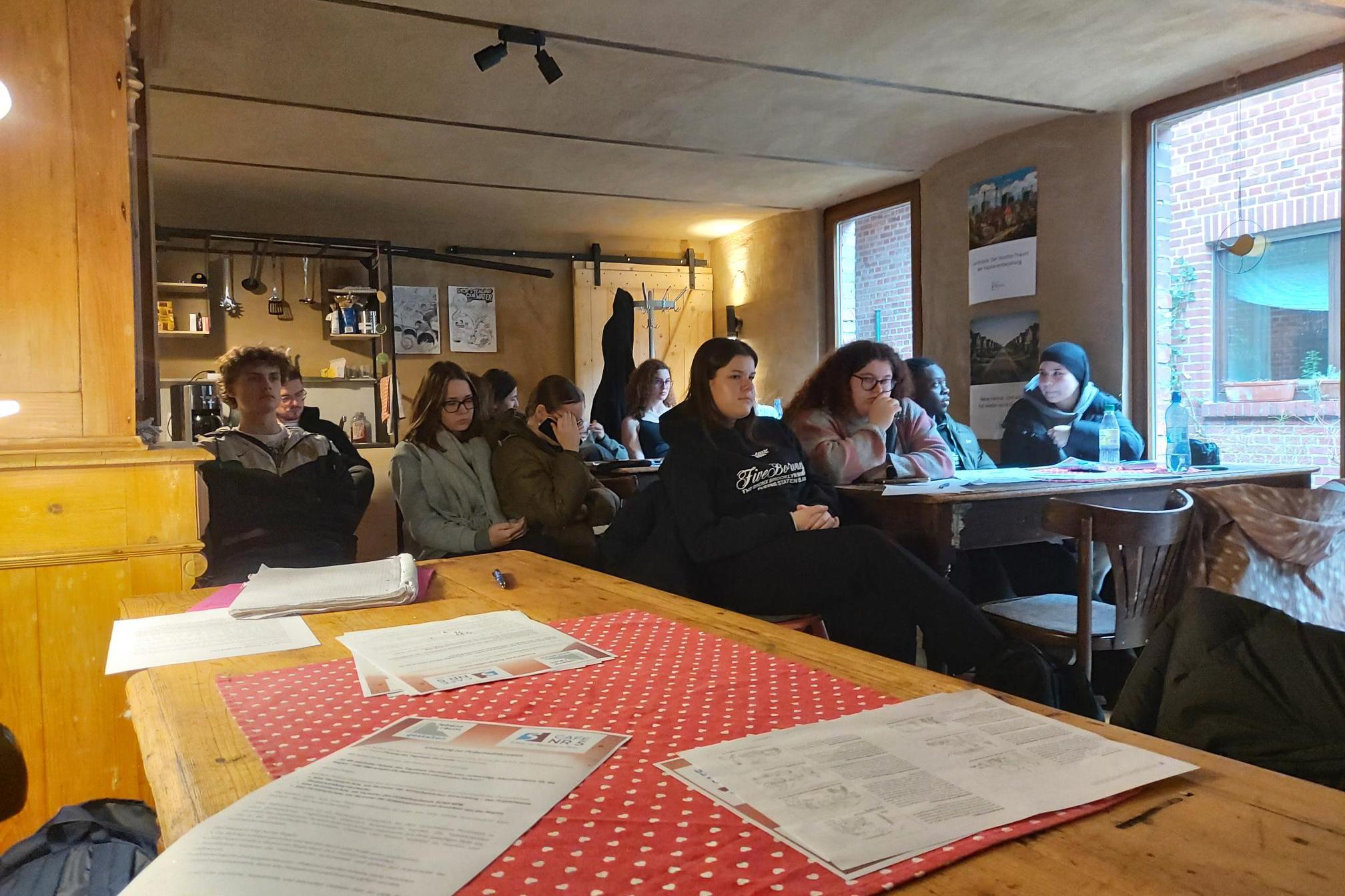 Mehrere Personen sitzen an Holztischen in einem Raum mit großen Fenstern und Deckenlampen, auf den Tischen liegen Papiere und ein rot-weiß gepunktetes Tischtuch.