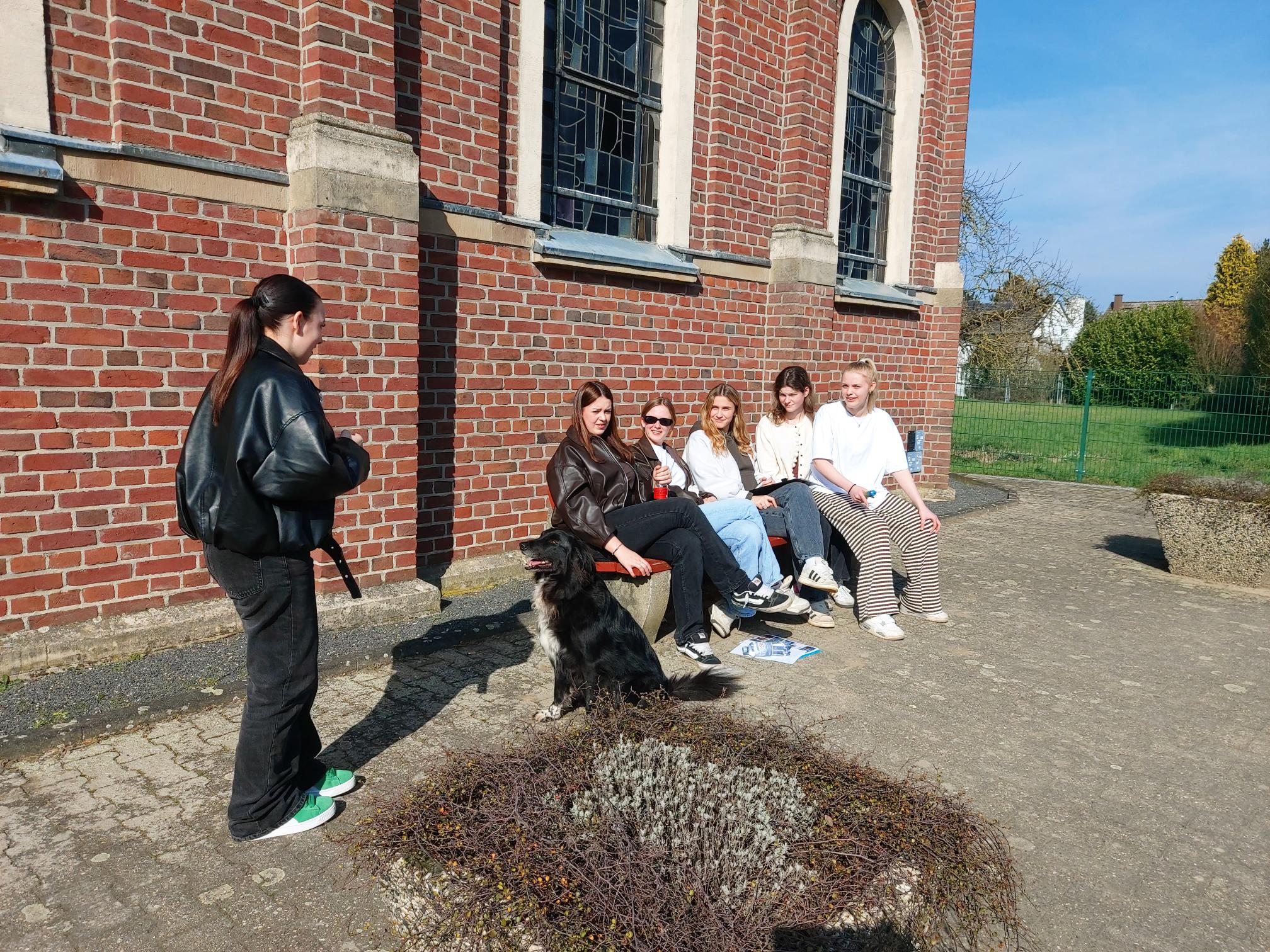 Gruppe von fünf Personen sitzt auf einer Bank vor einer Backsteinmauer mit Fenstern, eine Person steht daneben, ein Hund sitzt auf dem Boden davor