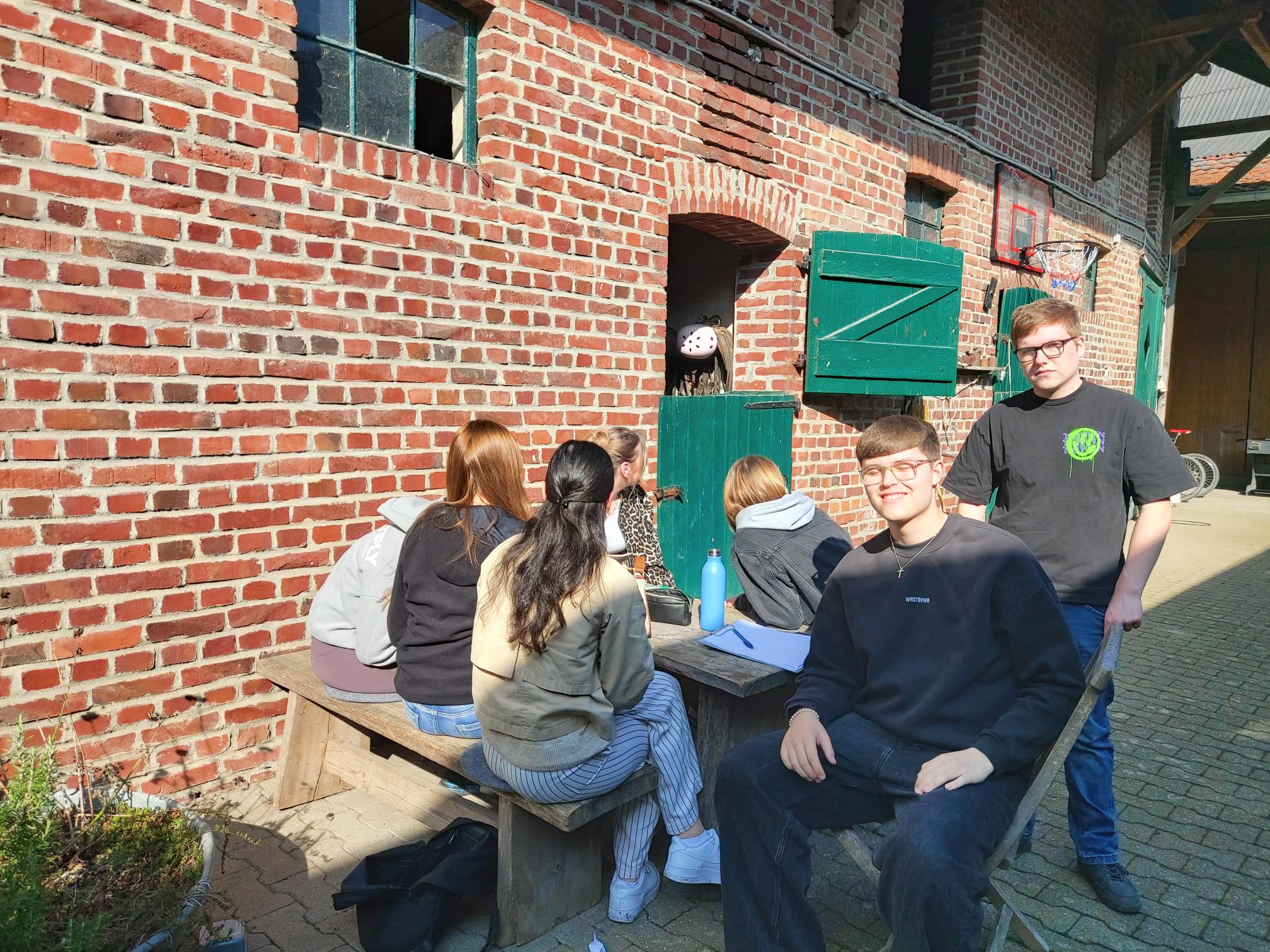 Gruppe von Jugendlichen sitzt an einem Holztisch vor einer roten Backsteinmauer mit geöffnetem grünem Fensterladen und Basketballkorb