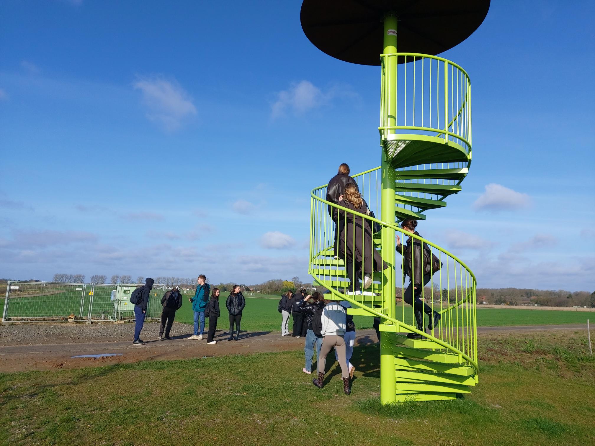 Gruppe von Menschen um eine grün lackierte Wendeltreppe im Freien auf einer Wiese bei klarem Himmel