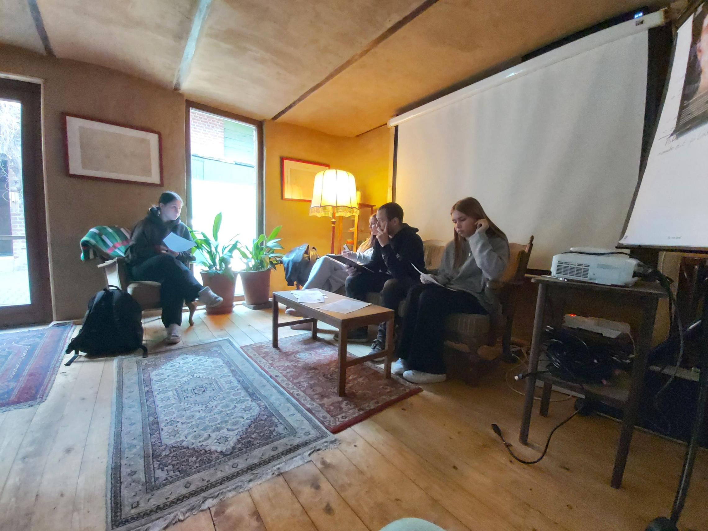 Vier Personen sitzen in einem Wohnzimmer auf Sesseln und einem Sofa, drei halten Tablets oder Smartphones, ein Tisch mit Pflanzen steht in der Mitte.