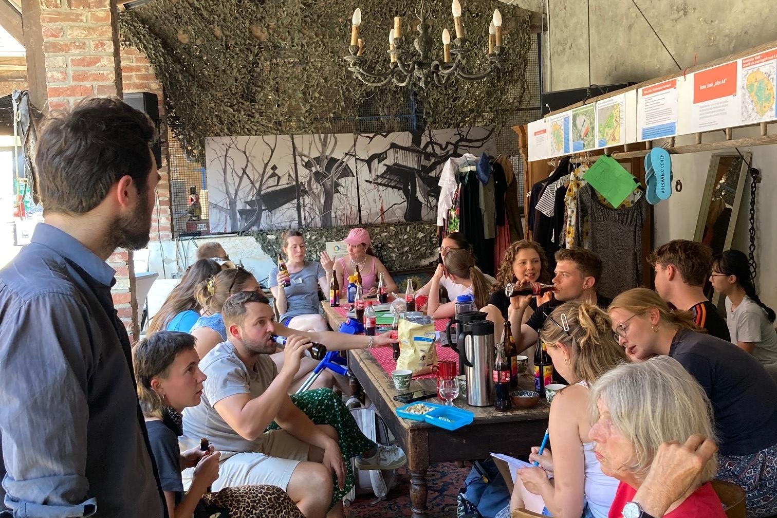 Gruppe von Menschen sitzt und steht um einen langen Tisch in einem überdachten Außenbereich, trinkt und unterhält sich