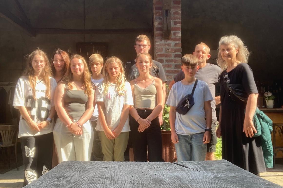 Gruppe von neun Personen steht vor einer Backsteinwand unter einem überdachten Bereich an einem sonnigen Tag