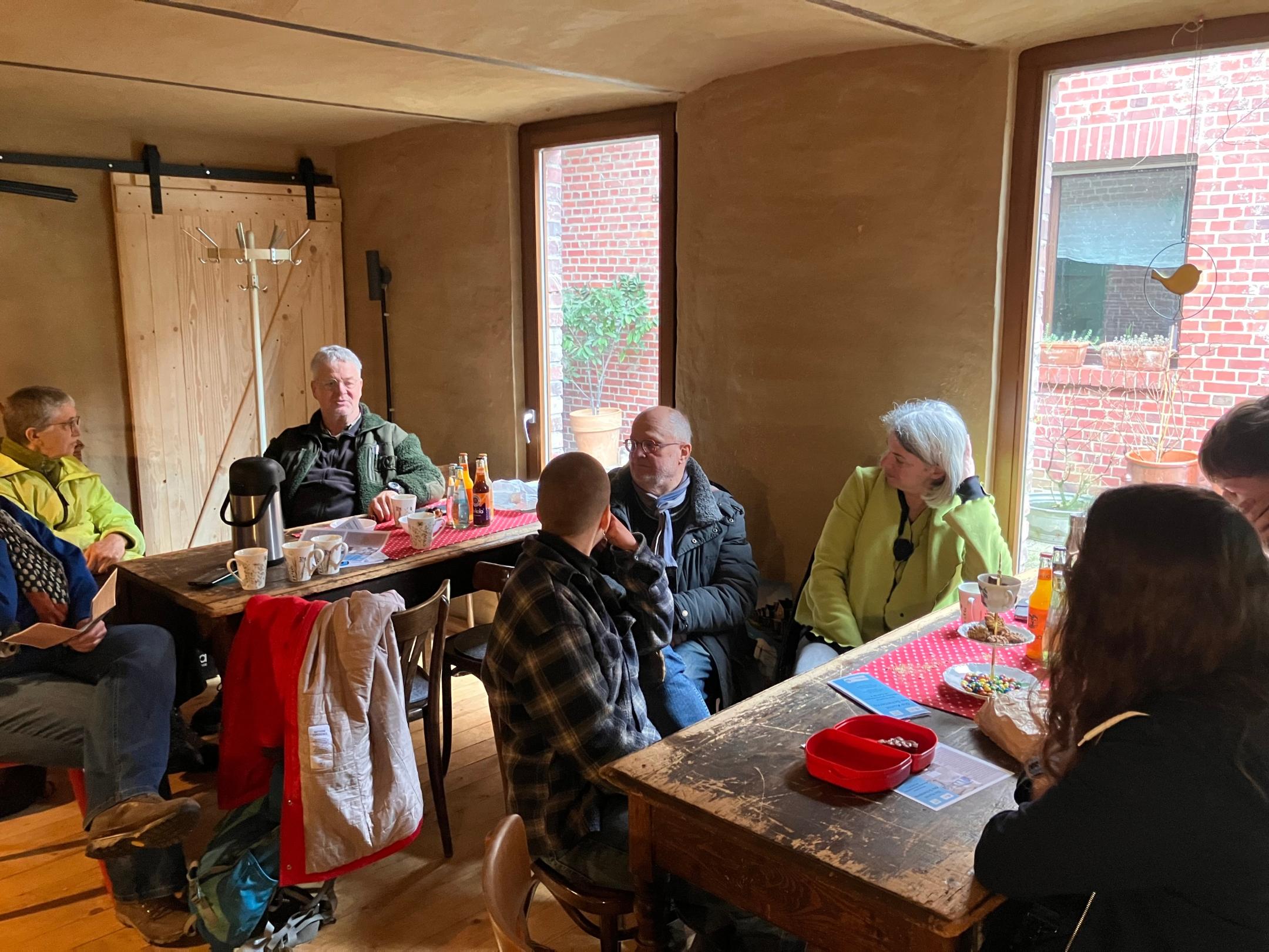 Gruppe von Menschen sitzt an zwei Holztischen in einem Raum mit Lehmwänden und großen Fenstern