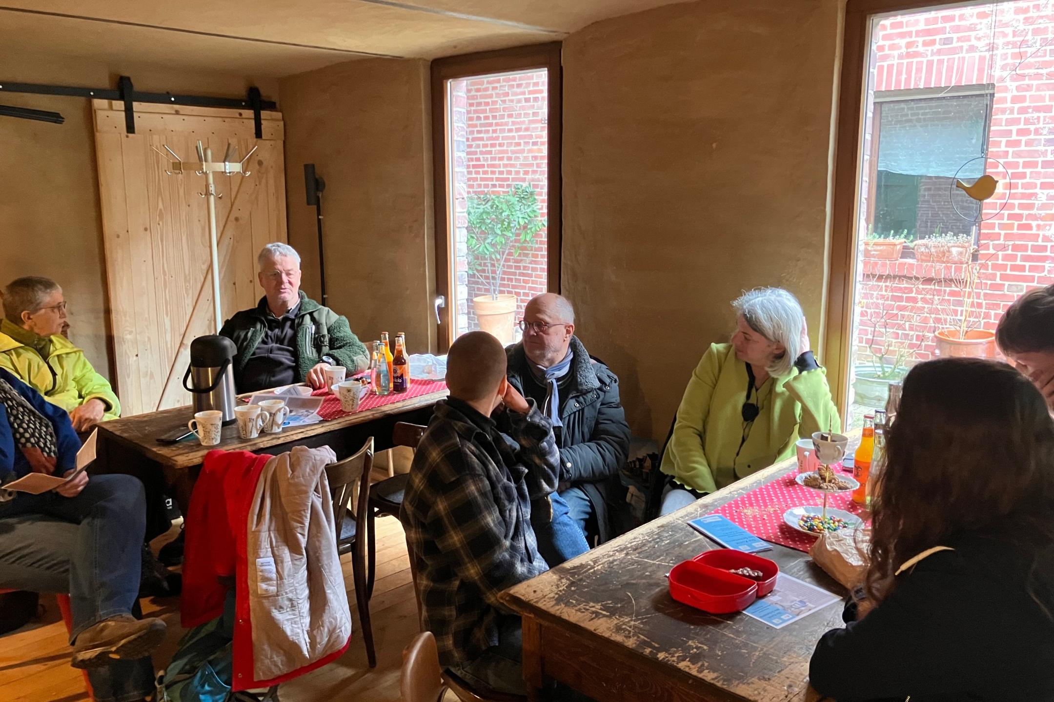 Gruppe von Menschen sitzt an zwei Holztischen in einem Raum mit Lehmwänden und großen Fenstern