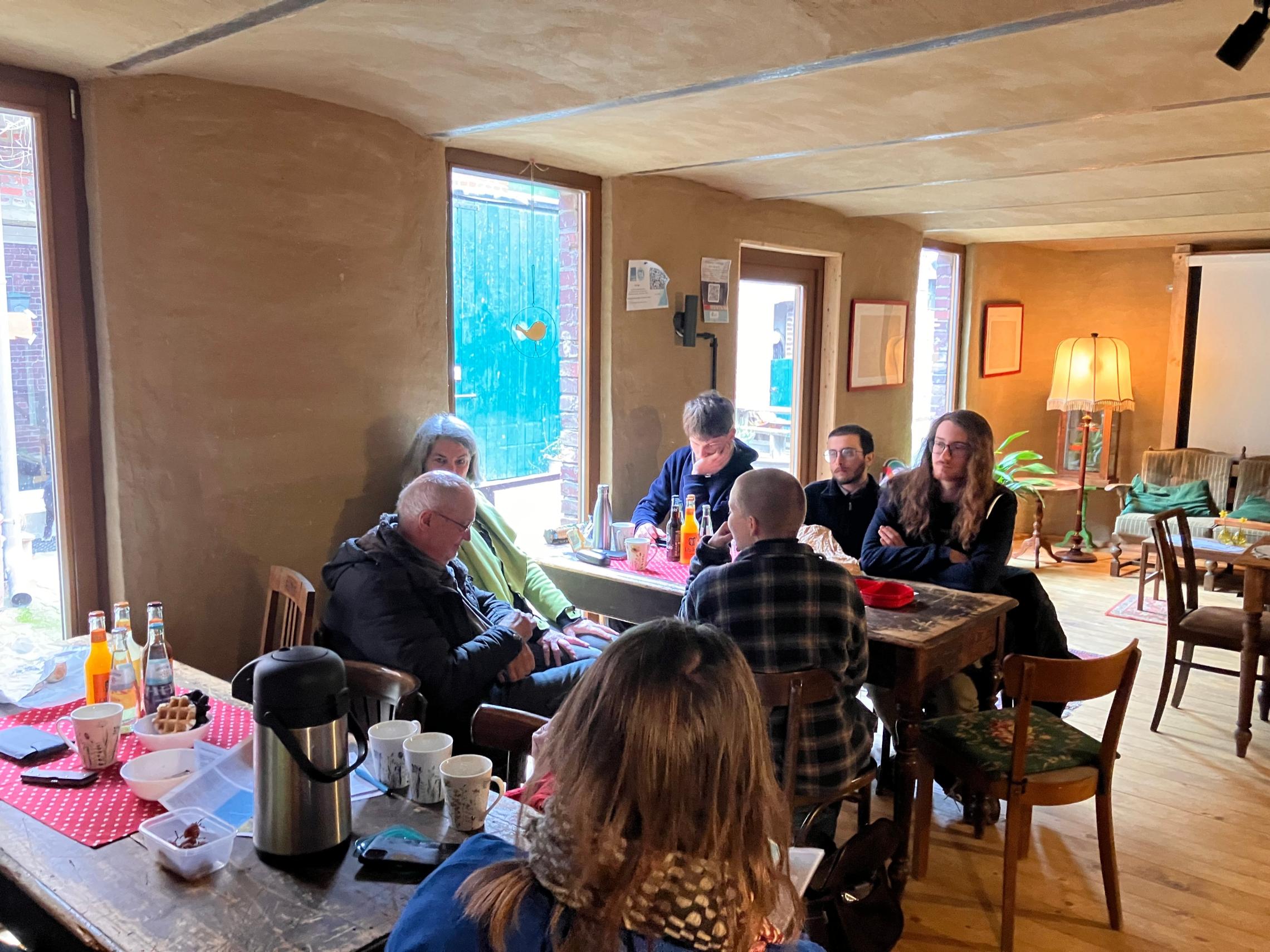 Gruppe von Menschen sitzt an zwei Holztischen in einem gemütlichen Raum mit Fenstern, Lampen und Stühlen