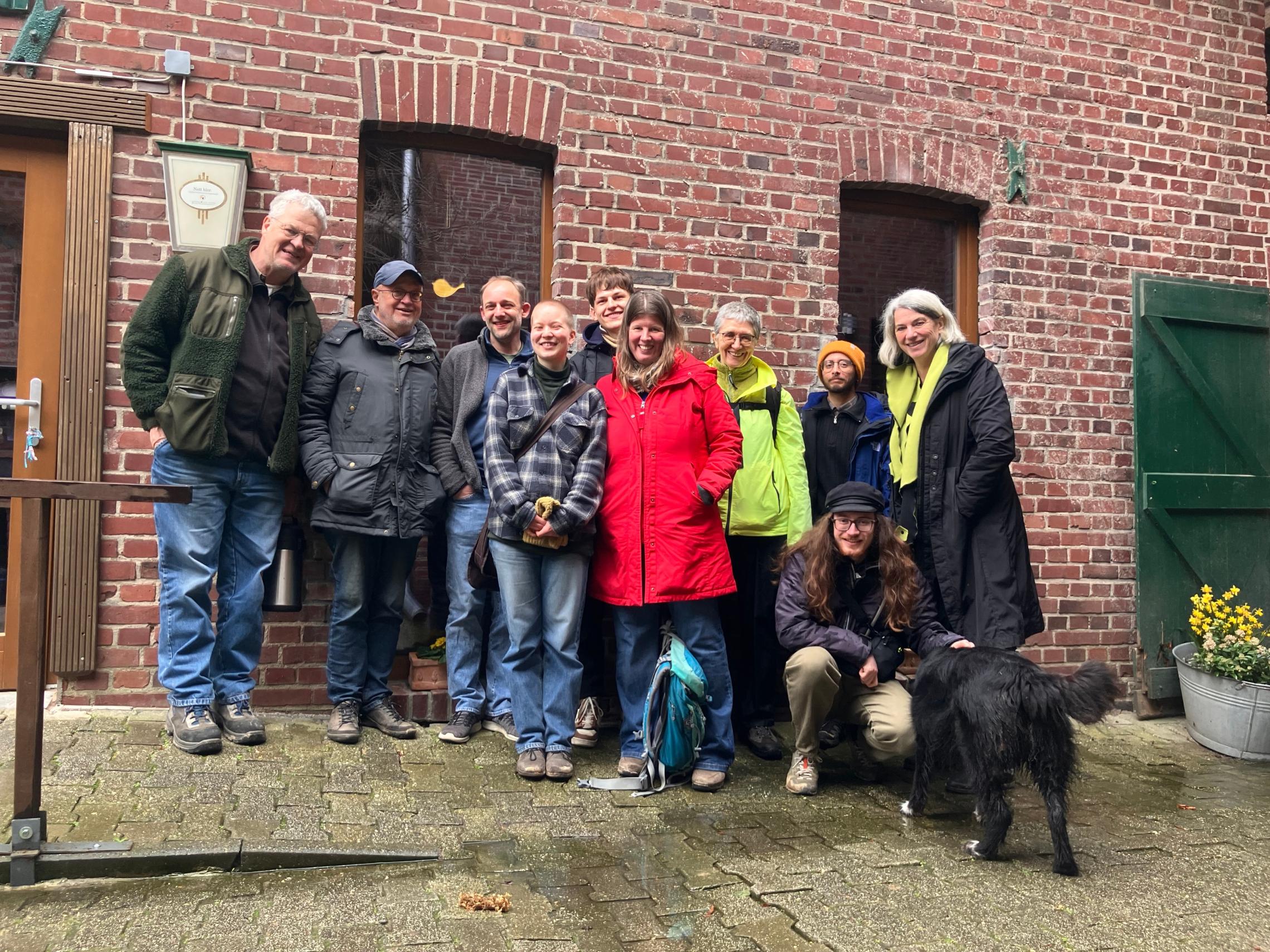 Gruppe von Menschen in Outdoor-Kleidung vor roter Backsteinwand mit zwei Fenstern und grünem Türflügel, eine Person hockt neben schwarzem Hund