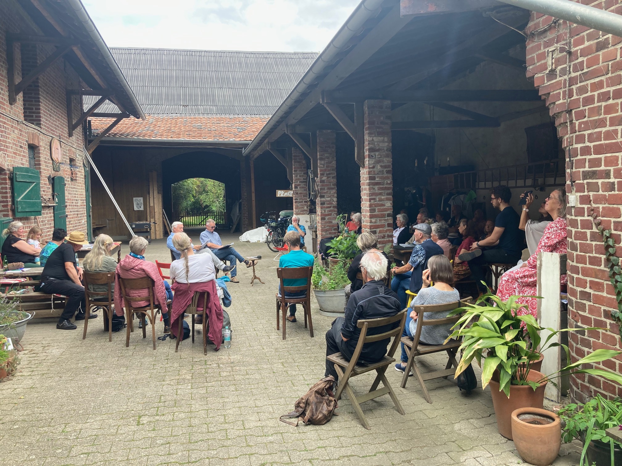 Gruppe von Menschen sitzt im Halbkreis auf Stühlen in einem Innenhof mit Backsteinmauern, einige Pflanzen sind sichtbar