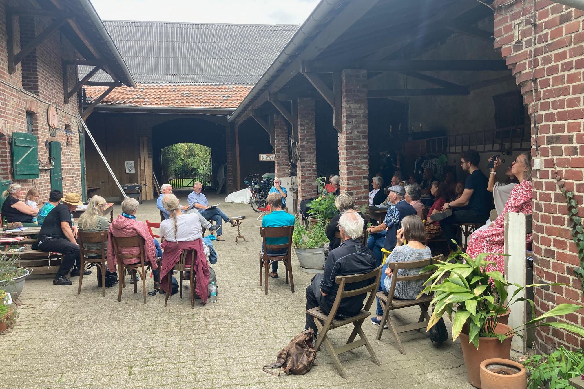 Gruppe von Menschen sitzt im Halbkreis auf Stühlen in einem Innenhof mit Backsteinmauern, einige Pflanzen sind sichtbar