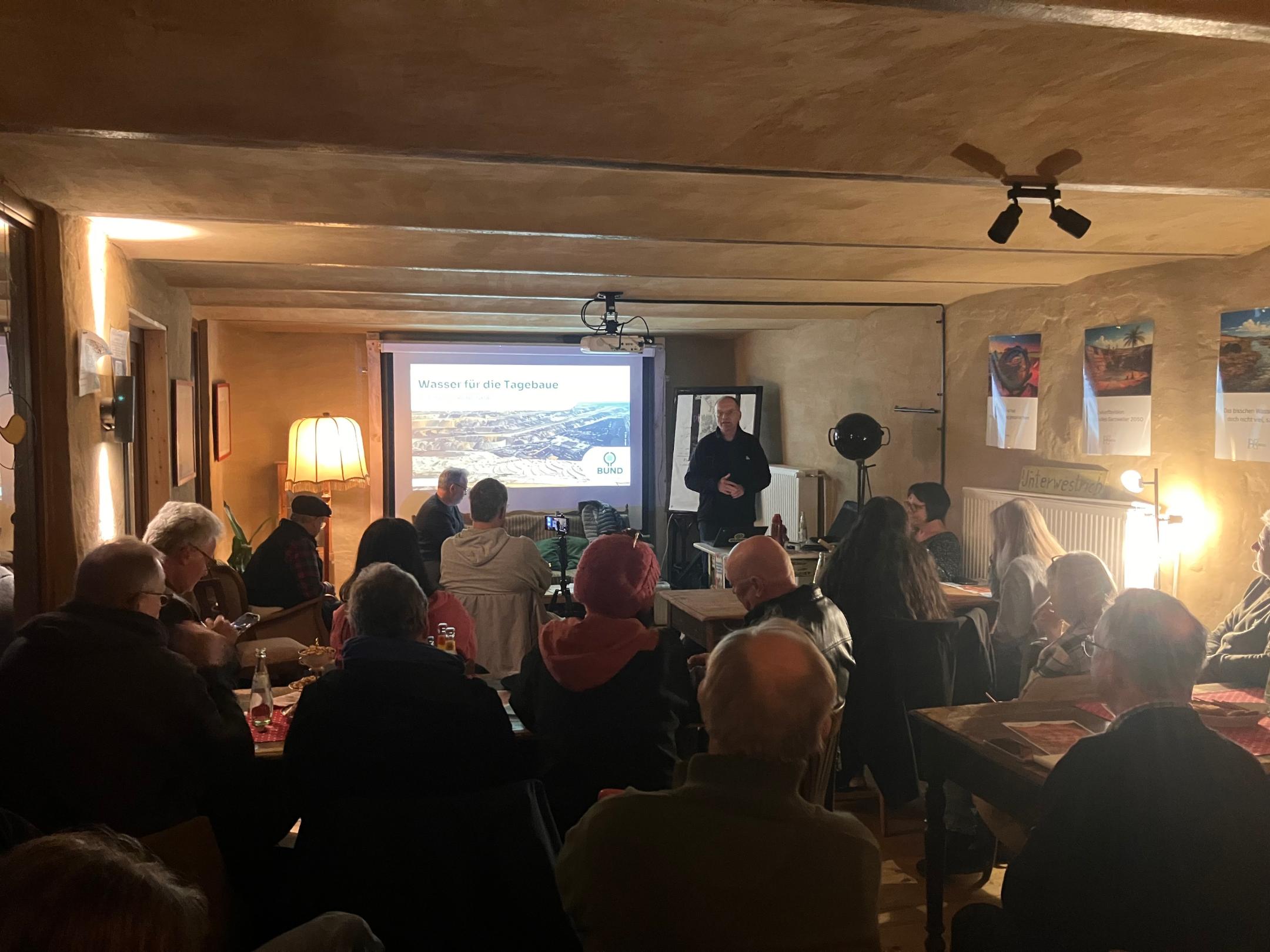 Mehrere Personen sitzen in einem Raum und hören einem Vortragenden zu, der vor einer Leinwand mit der Überschrift 'Wasser für die Tagebaue' steht