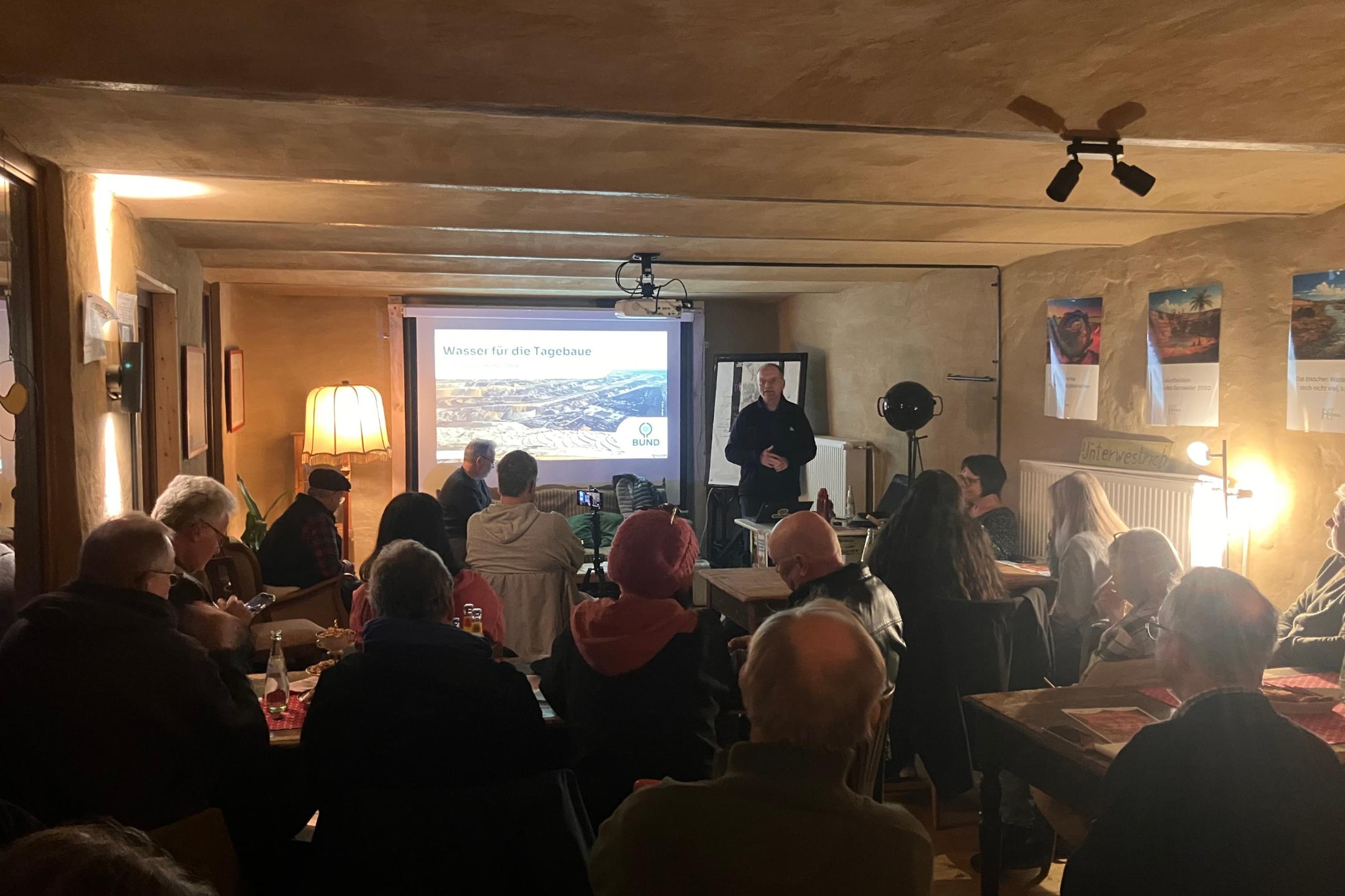 Mehrere Personen sitzen in einem Raum und hören einem Vortragenden zu, der vor einer Leinwand mit der Überschrift 'Wasser für die Tagebaue' steht