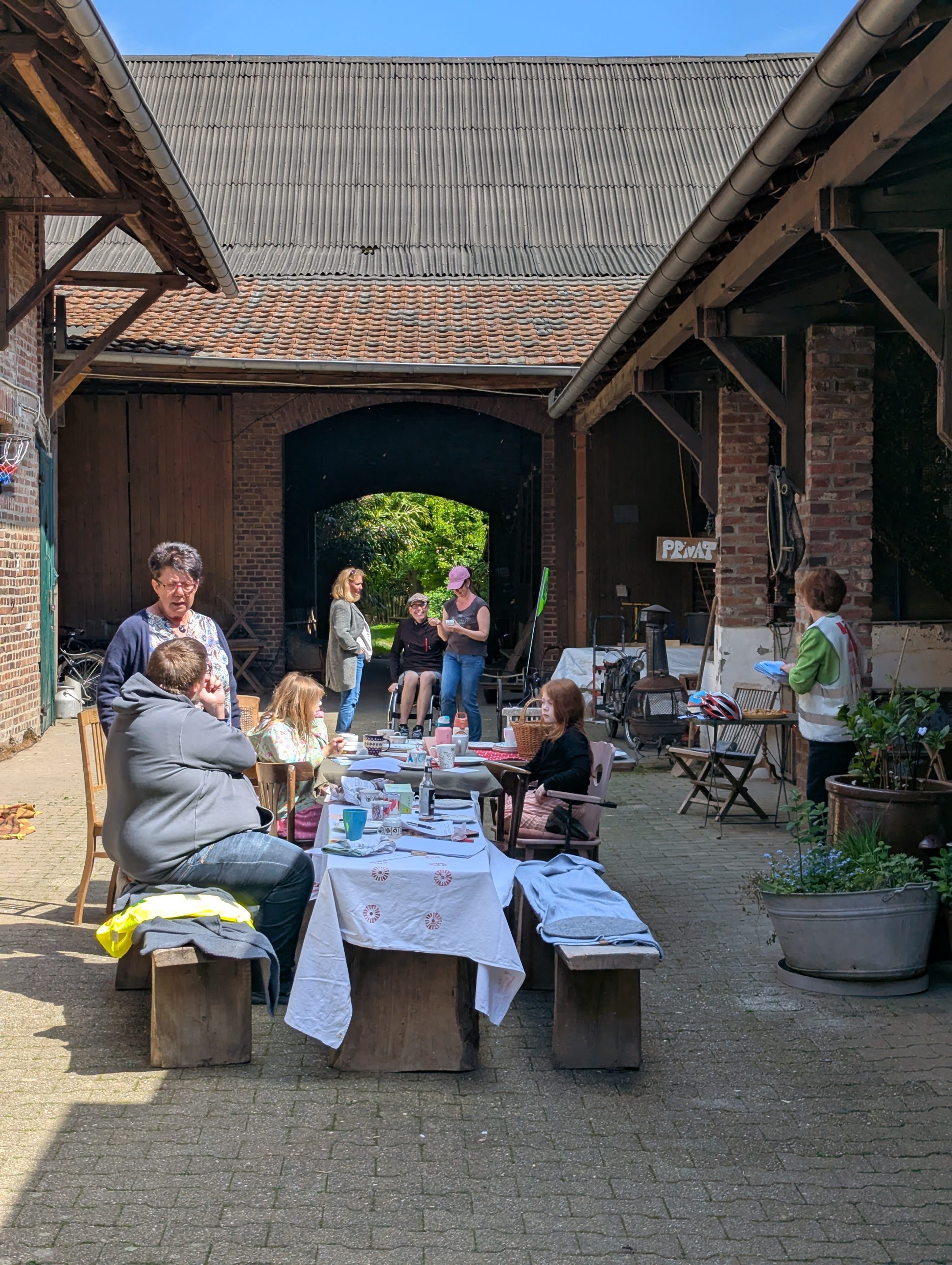Mehrere Personen sitzen und stehen an einem langen Tisch mit weißer Tischdecke in einem Innenhof zwischen zwei Gebäuden mit sichtbaren Holzbalken und Ziegeldächern