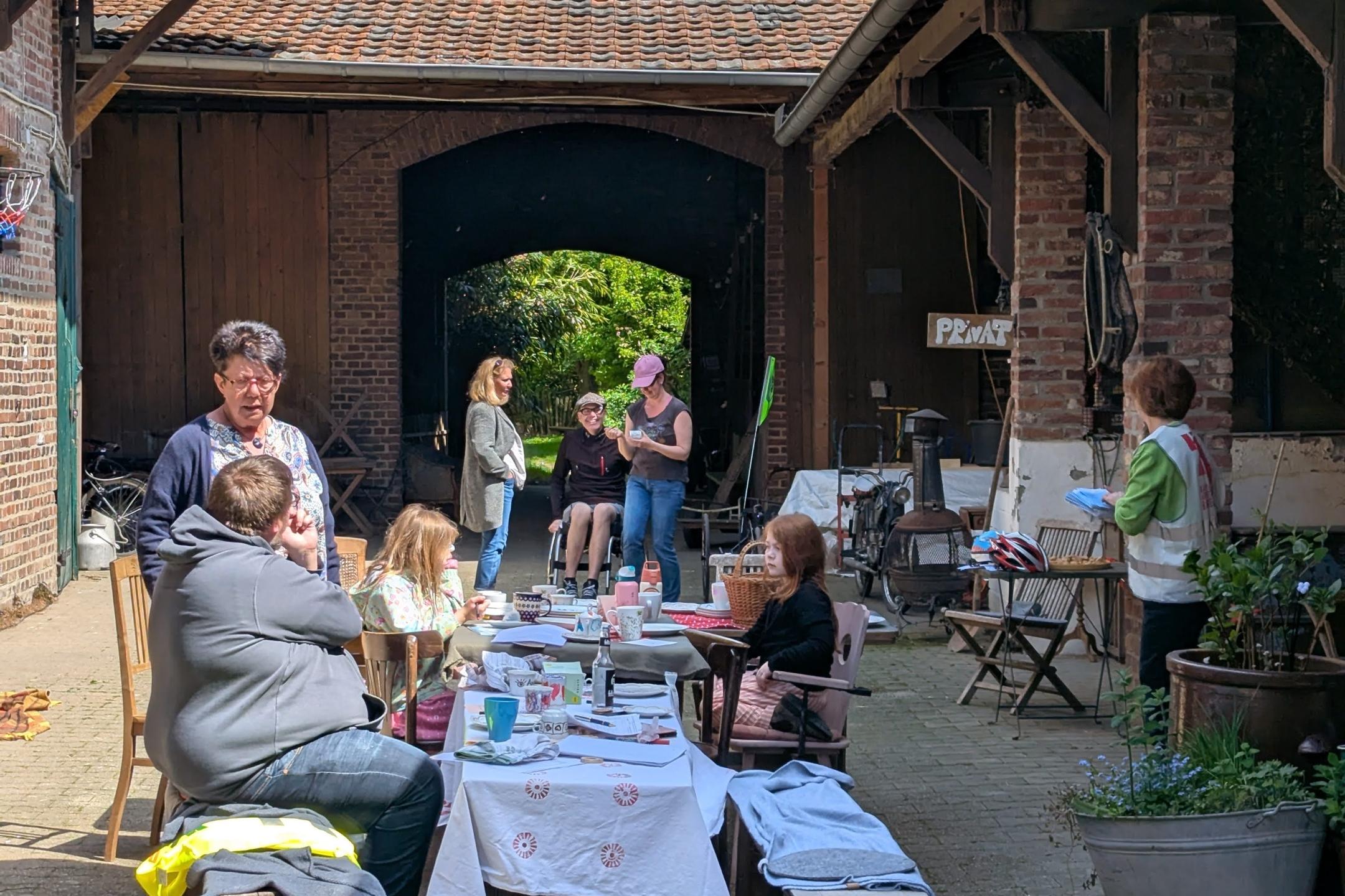 Mehrere Personen sitzen und stehen an einem langen Tisch mit weißer Tischdecke in einem Innenhof zwischen zwei Gebäuden mit sichtbaren Holzbalken und Ziegeldächern